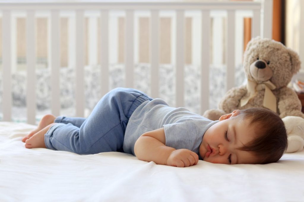 Baby sleeping in their crib with a stuffed bear.
