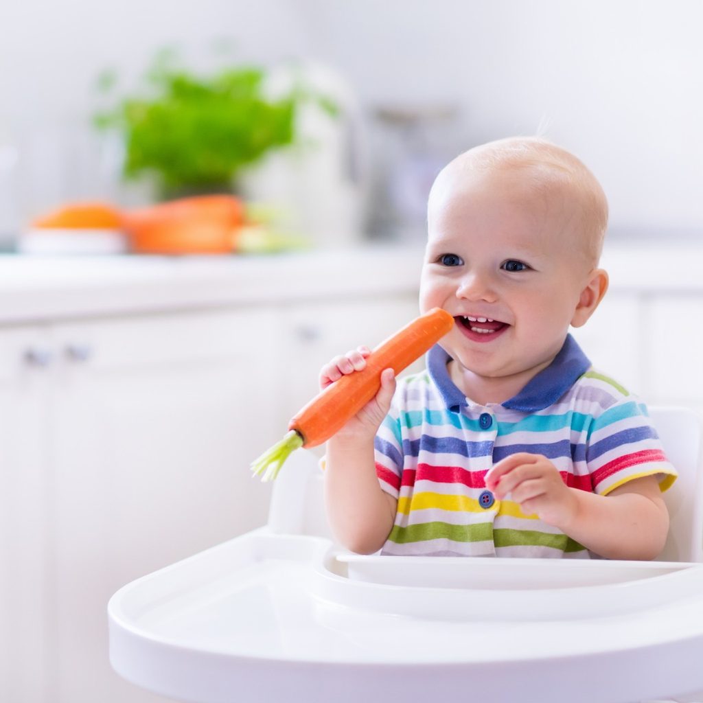 Smiling baby in high chair holding a carrot