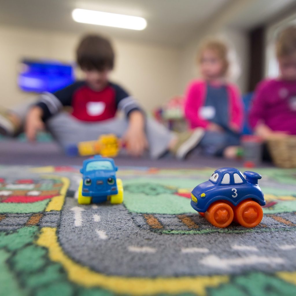 Three kids at daycare in background with street-themed rug and cars in foreground