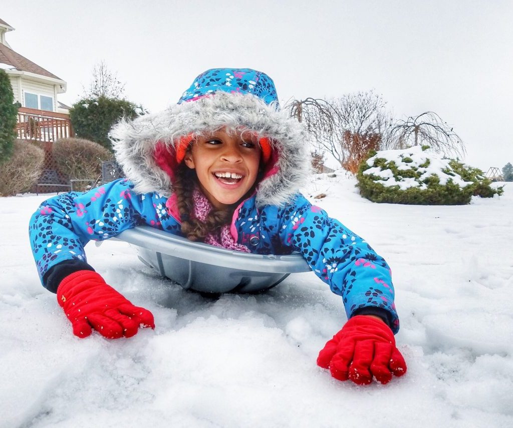 A child playing in snow all bundled up sliding on a sled