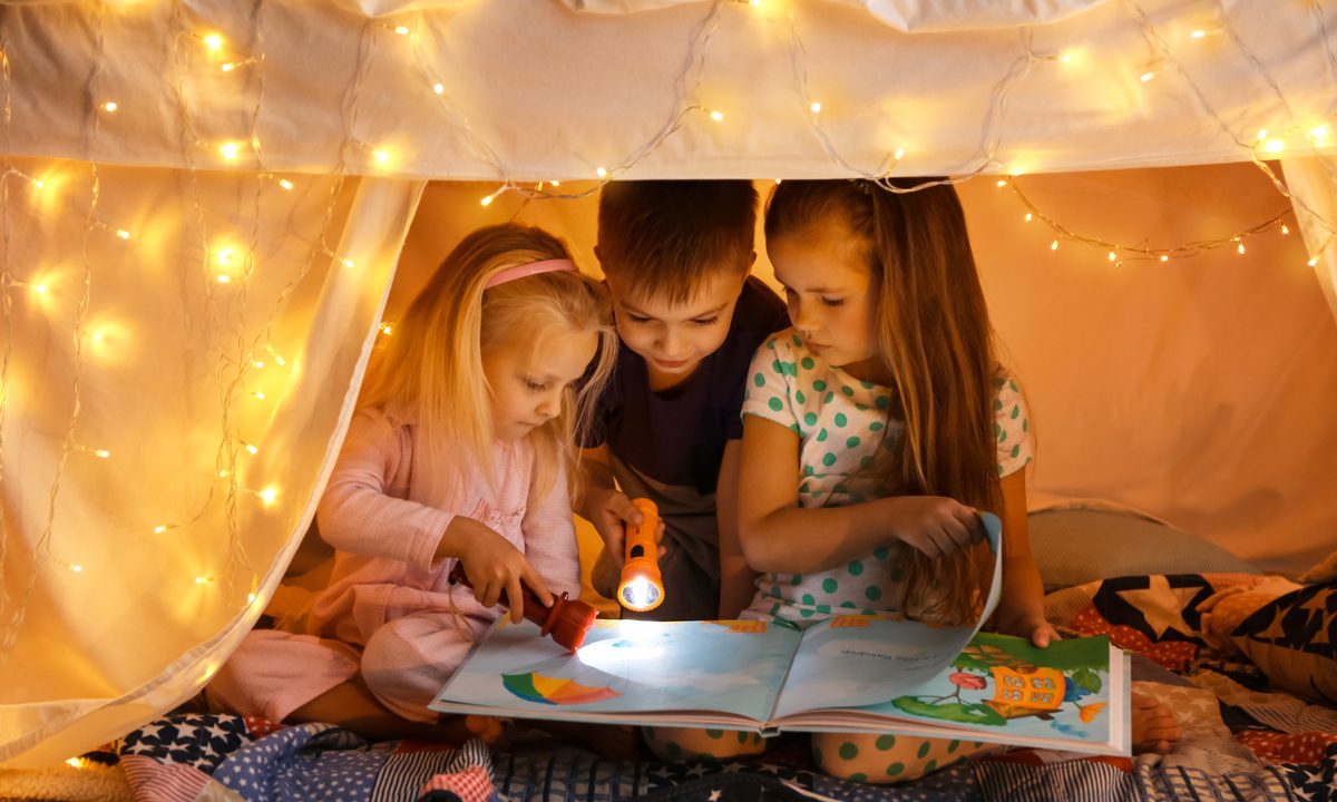Three kids reading a book inside of a blanket fort