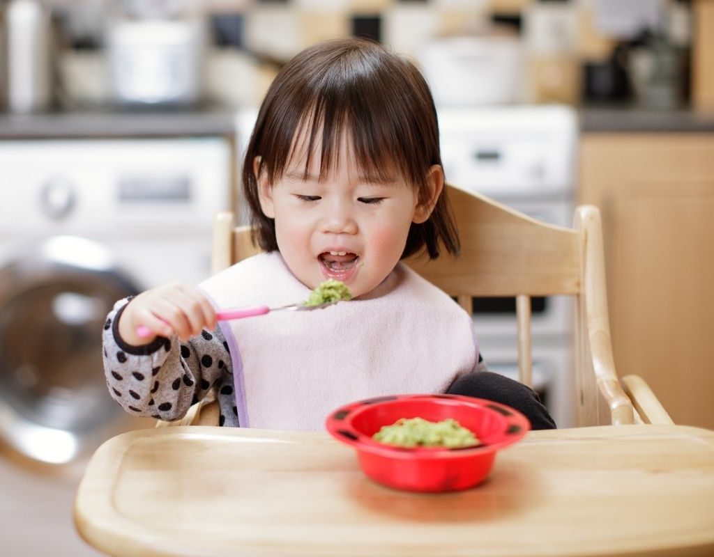 Baby in highchair eating avocado puree.