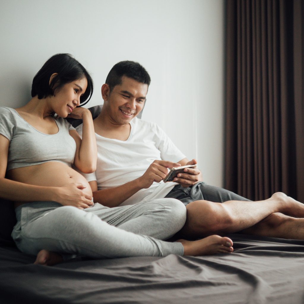 Man and pregnant woman sit on bed looking at a smartphone