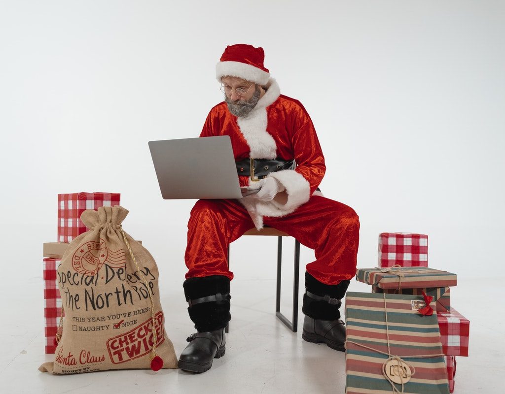 Santa looking at list on laptop with presents around him.