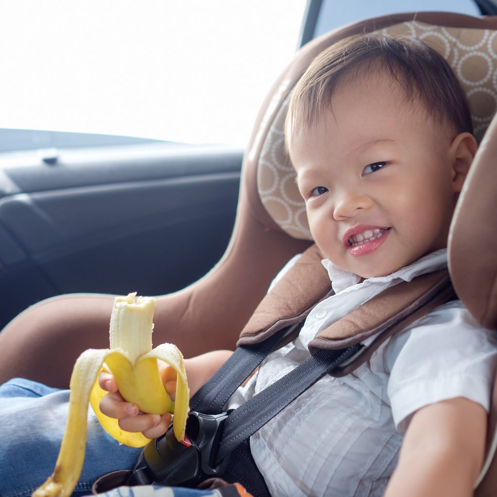 Toddler boy eating a banana in his car seat