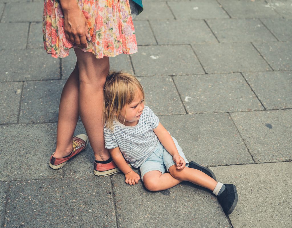 A toddler crying on the ground while leaning against his mother's legs.