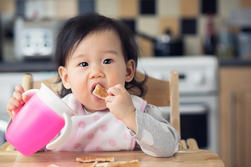 A young toddler eating food and holding a sippy cup.