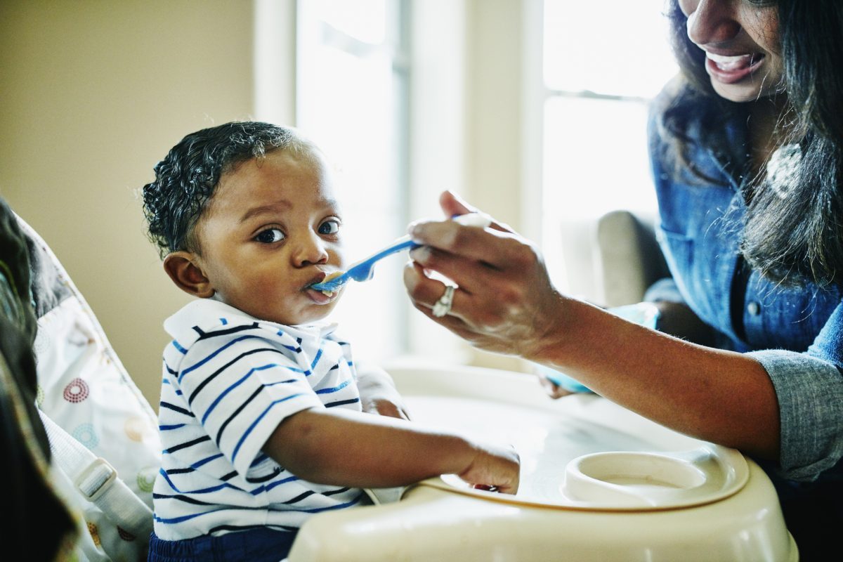 A parent feeding their infant baby food while the baby sits in a high chair