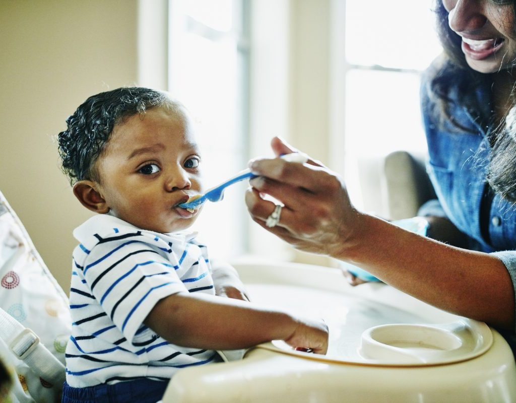 A parent feeding their infant baby food while the baby sits in a high chair