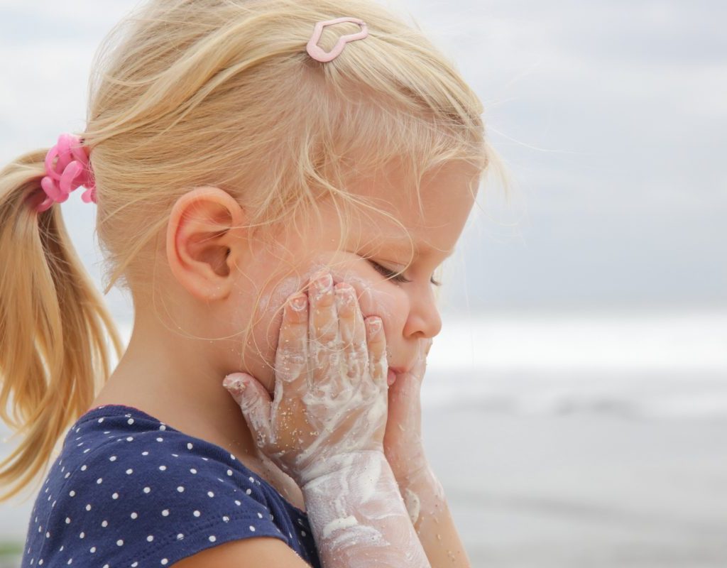 A little girl has sunscreen on her arms as she applys more to her face.