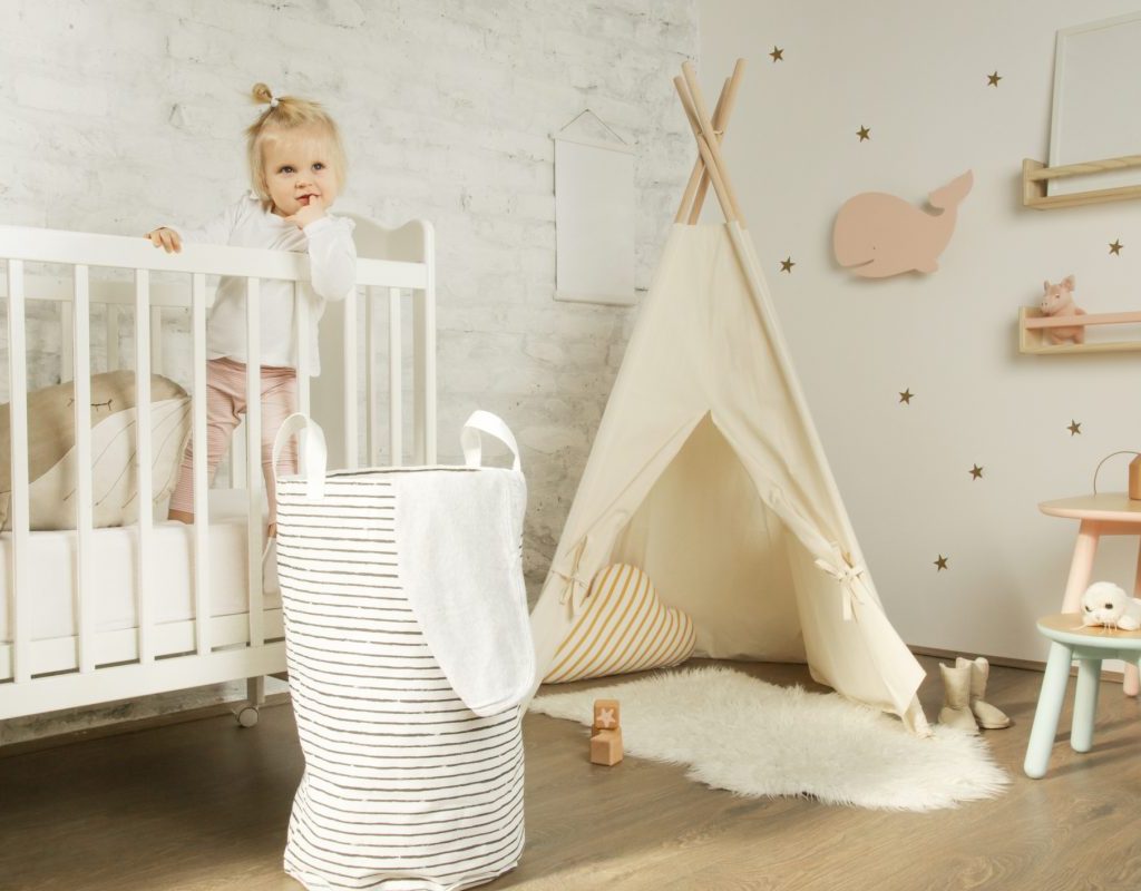 A little girl in her crib in her room with a laundry basket by her crib
