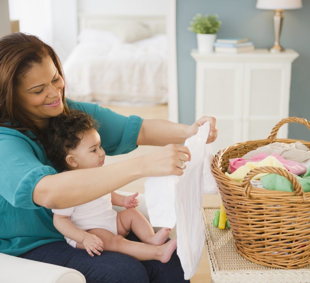 A mother doing laundry with her baby in her lap and the laundry basket sitting in front of her