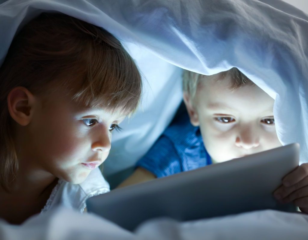 Two kids watching a tablet under a blanket.