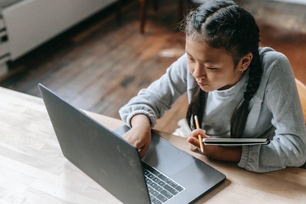 A child sitting at a desk while holding a notebook and typing on the laptop.