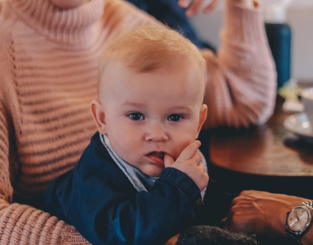 A baby sitting in his parent's lap while chewing on his thumb.