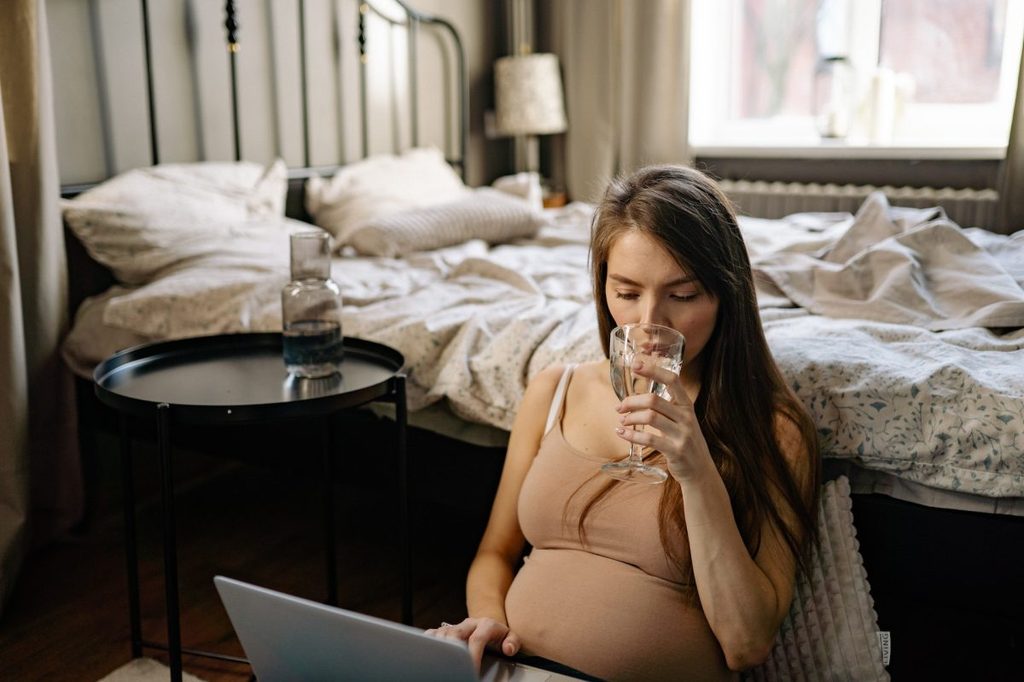 A pregnant woman sitting on the floor drinking a glass of water.