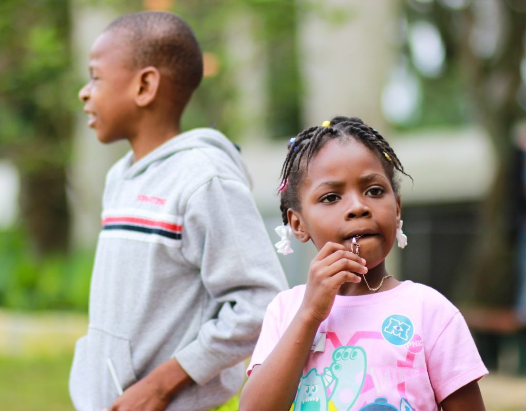 A child is chewing on their necklace with a sibling in the background.