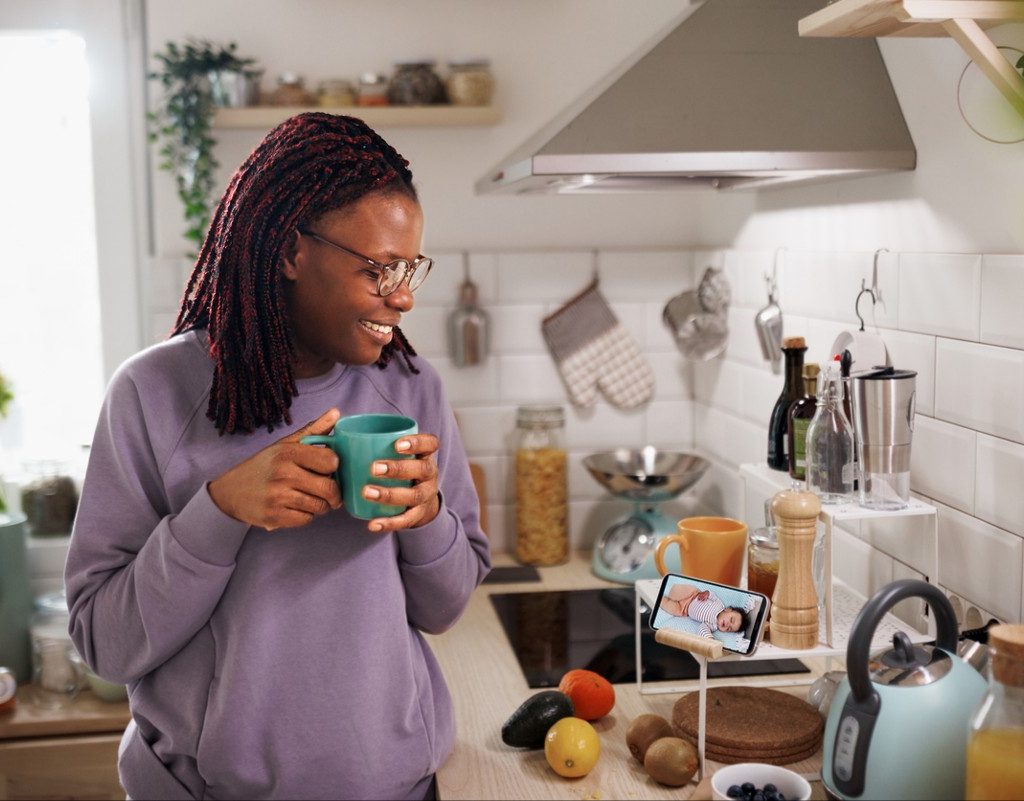 Smiling woman having coffee in kitchen using phone to monitor her sleeping baby