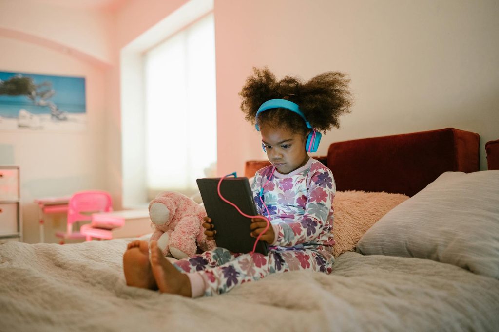 A child sitting on a bed, on a tablet with headphones on.
