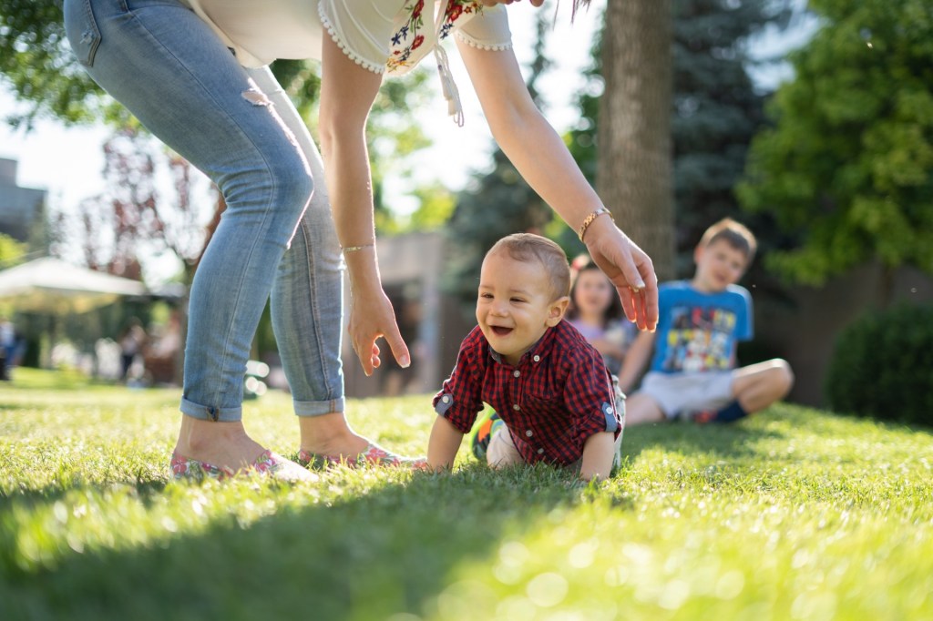 Toddler crawls on grass in park with mother watching.