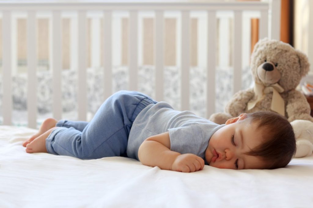 Baby sleeping in their crib with a stuffed bear.