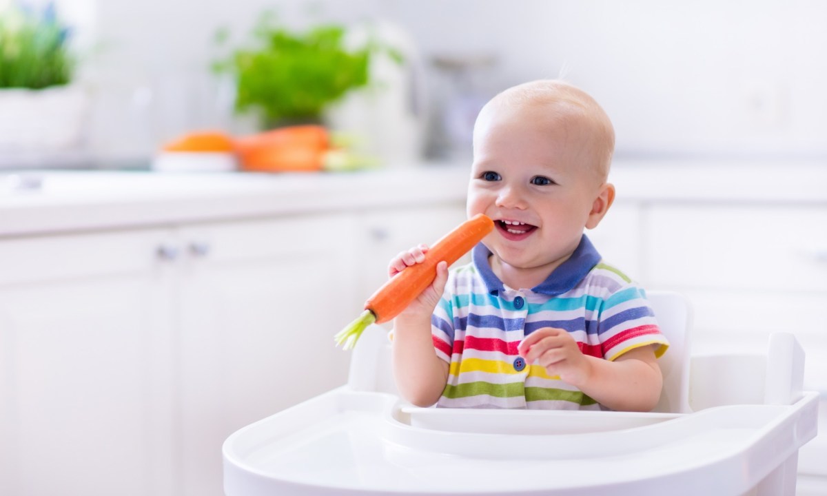 Smiling baby in high chair holding a carrot