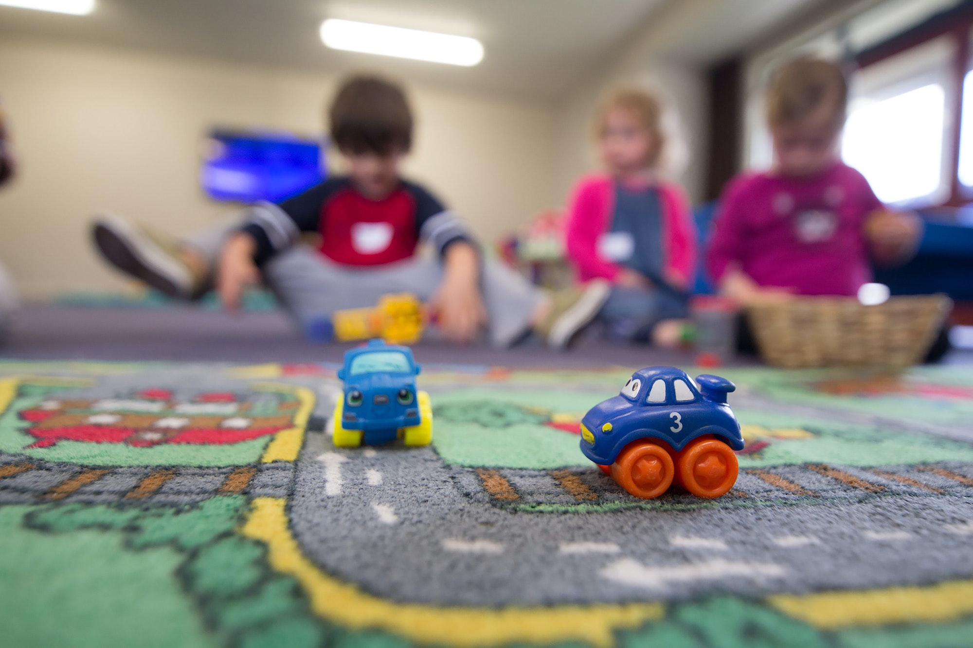Three kids at daycare in background with street-themed rug and cars in foreground