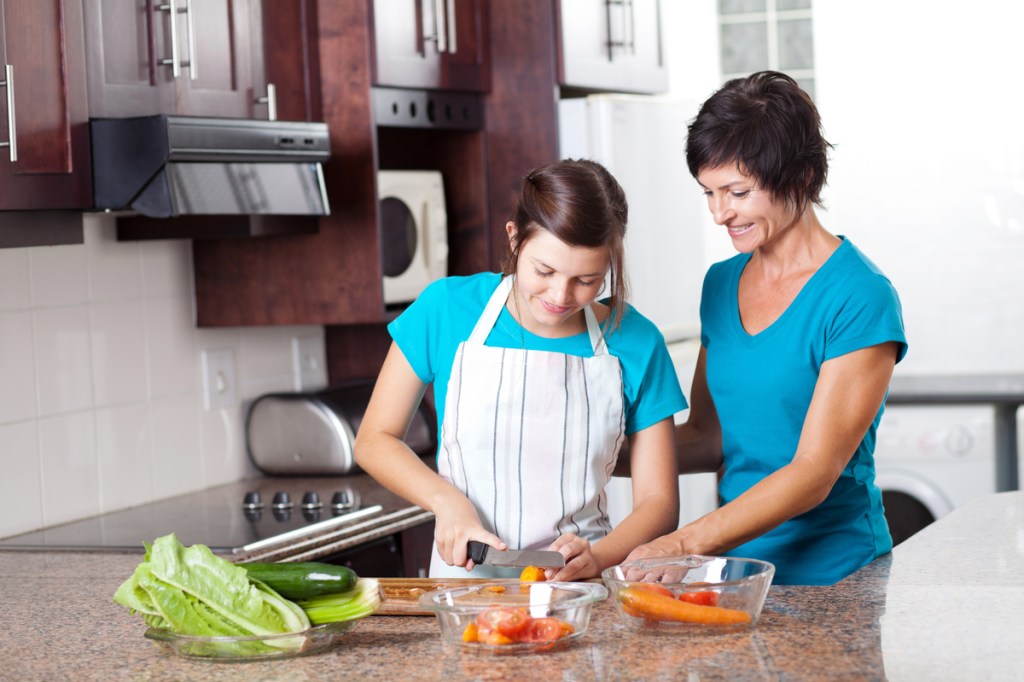 A teen chopping up vegetables while her parent watches.