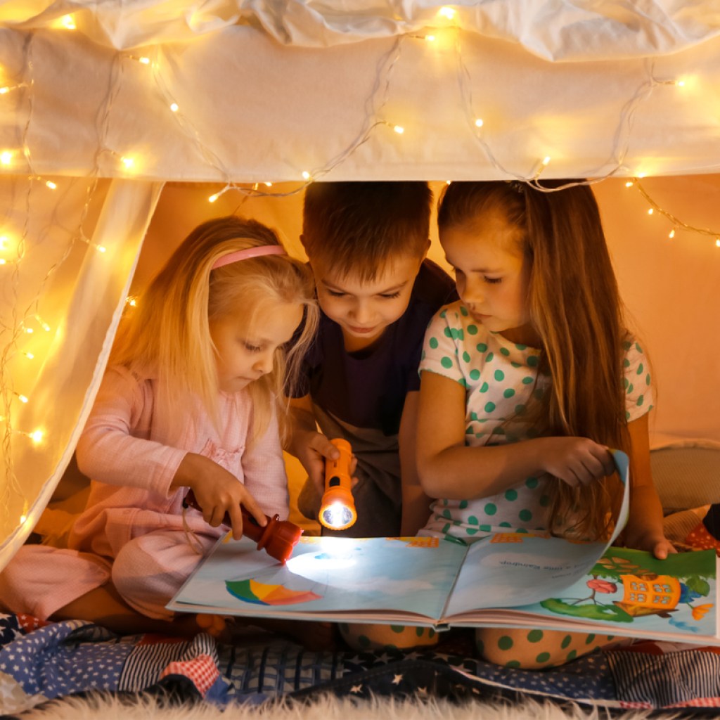Three kids reading a book inside of a blanket fort