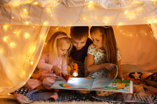 Three kids reading a book inside of a blanket fort