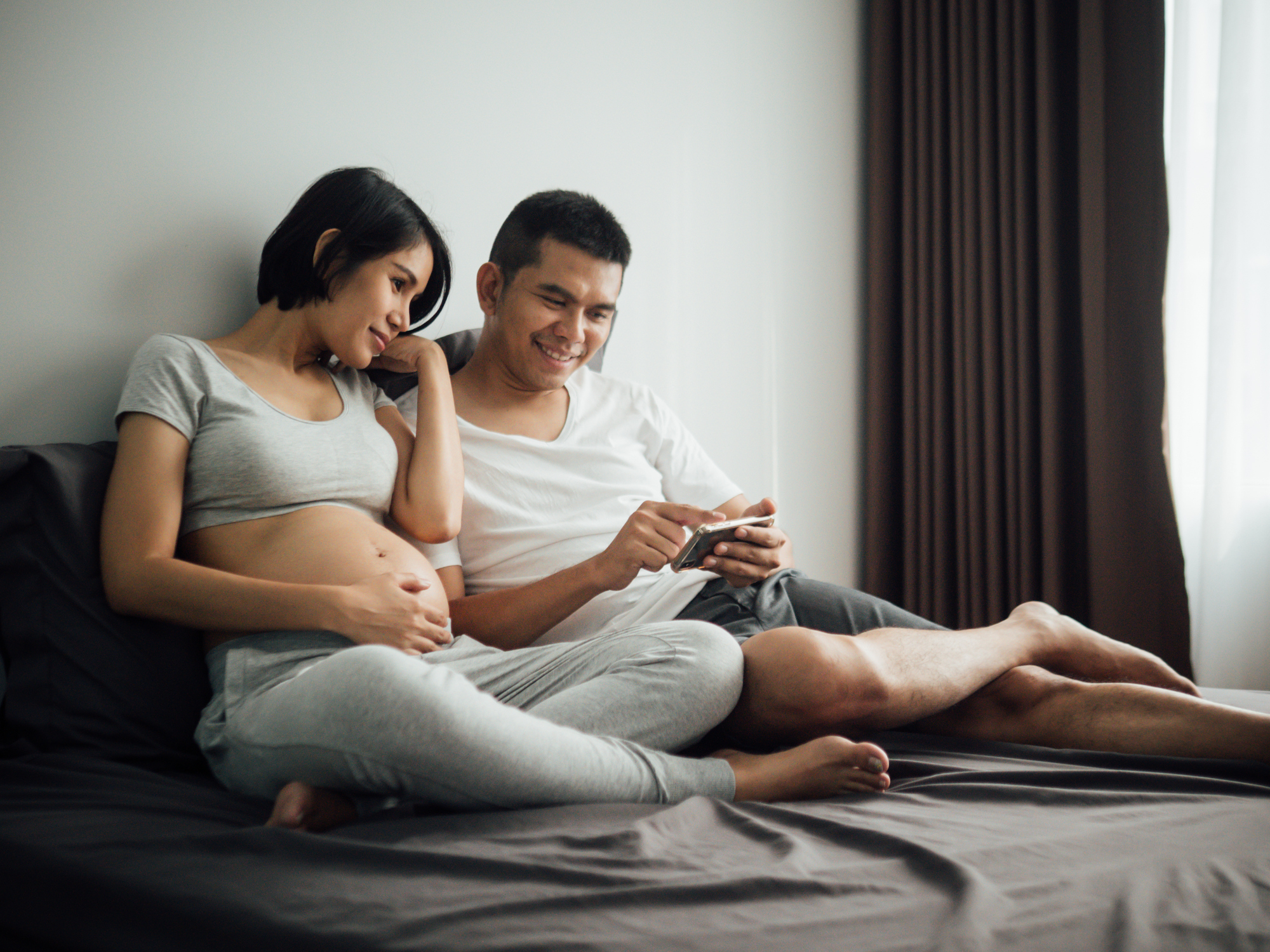Man and pregnant woman sit on bed looking at a smartphone