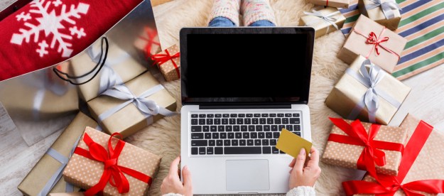A person doing holiday shopping on their laptop with presents all around them.