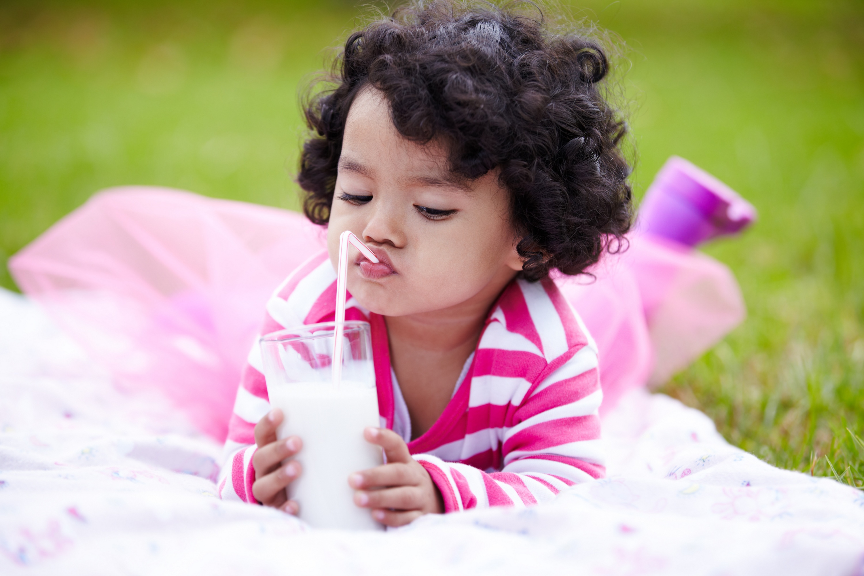 Toddler girl drinking milk through a straw while lying on the grass