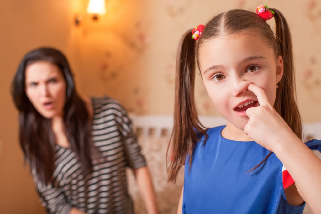 A young girl picking her nose with her mother in the background looking grossed out.