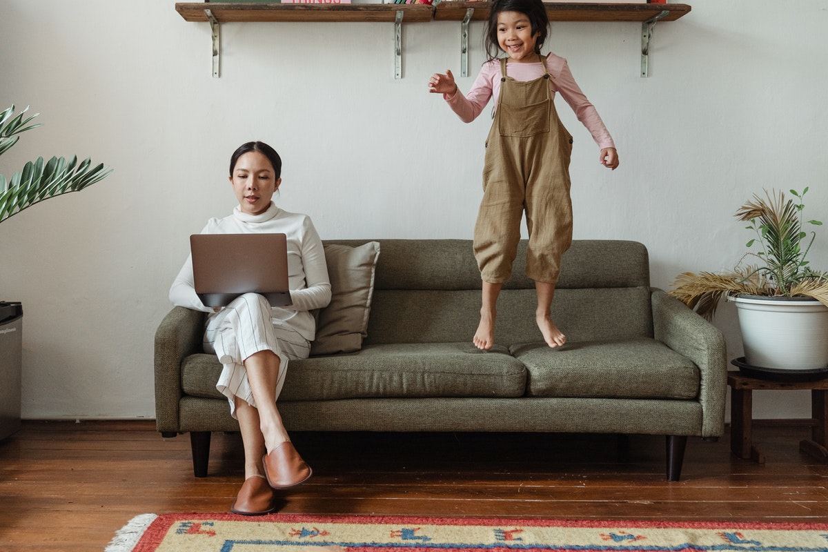 Parent working while child jumps on couch beside her.