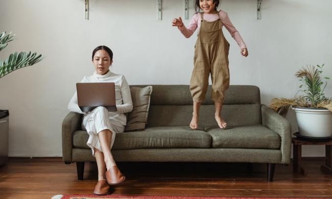 Parent working while child jumps on couch beside her.
