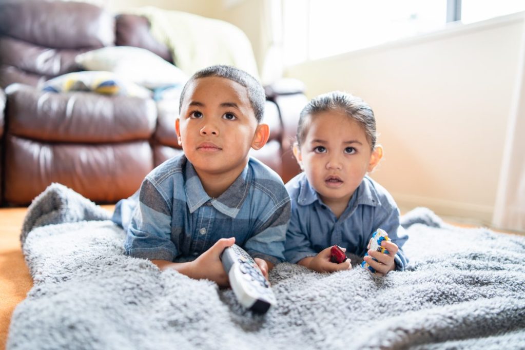 Two siblings lying on the floor watching tv together.