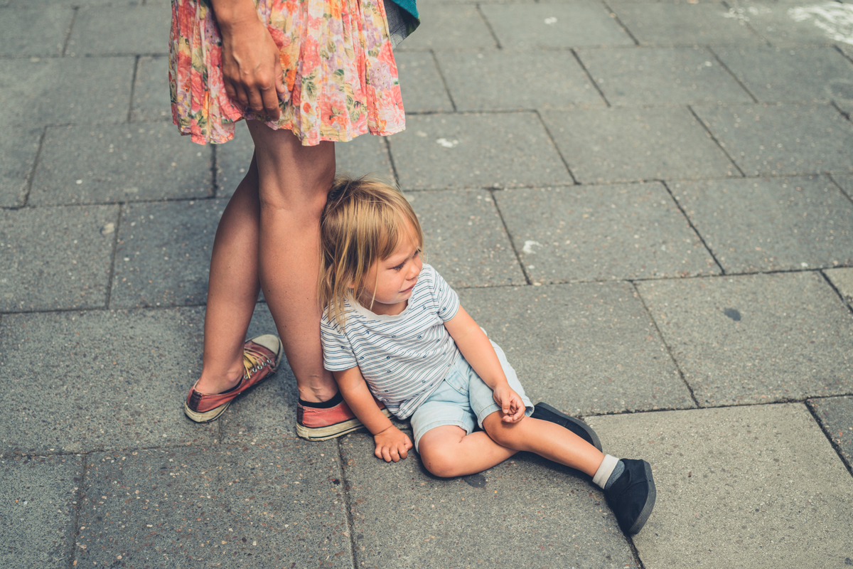 A toddler crying on the ground while leaning against his mother's legs.