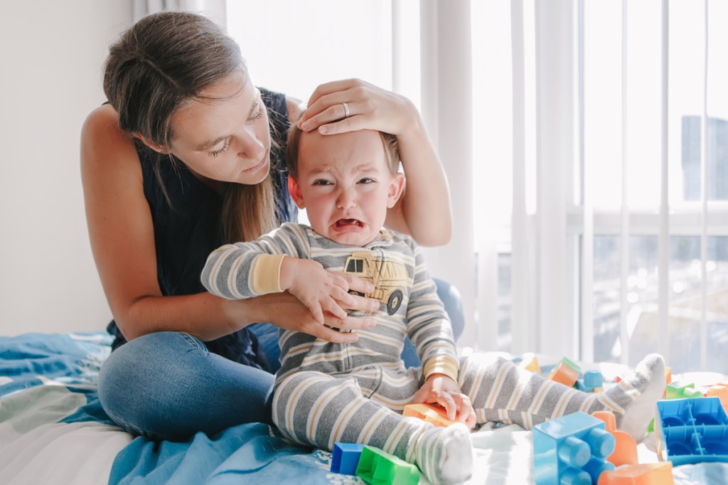 A mother holding her crying toddler sitting on the bed.