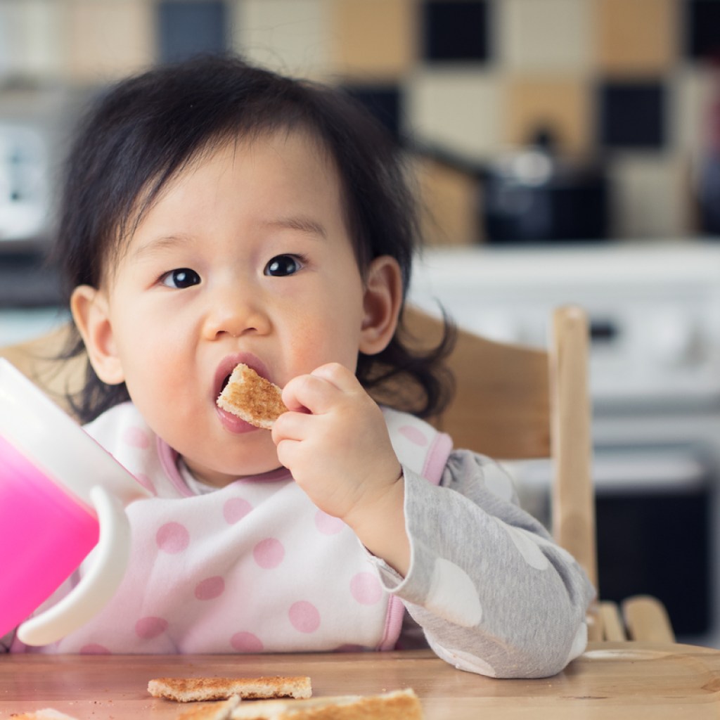 A young toddler eating food and holding a sippy cup.