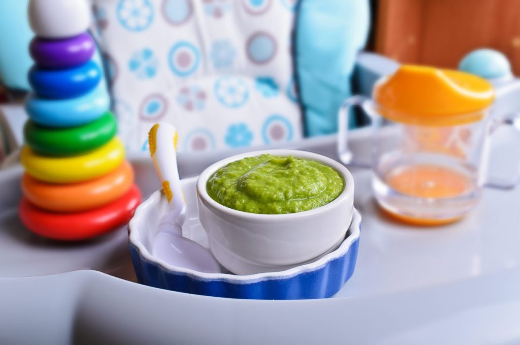 A highchair set up with a baby toy, sippy cup, and green baby food.