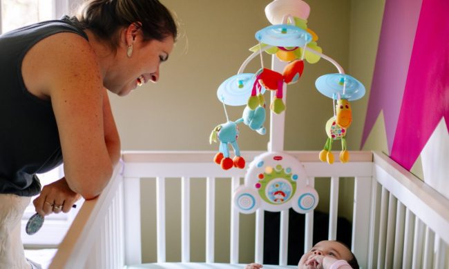A mother looking at her baby in the crib with a baby mobile.