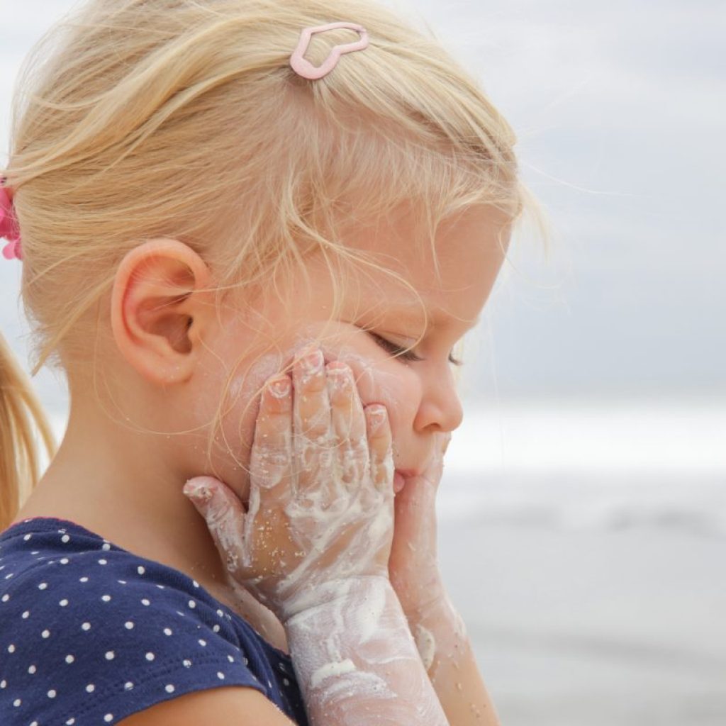 A little girl has sunscreen on her arms as she applys more to her face.