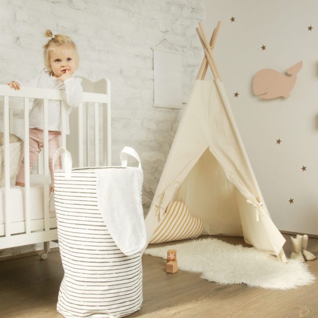 A little girl in her crib in her room with a laundry basket by her crib