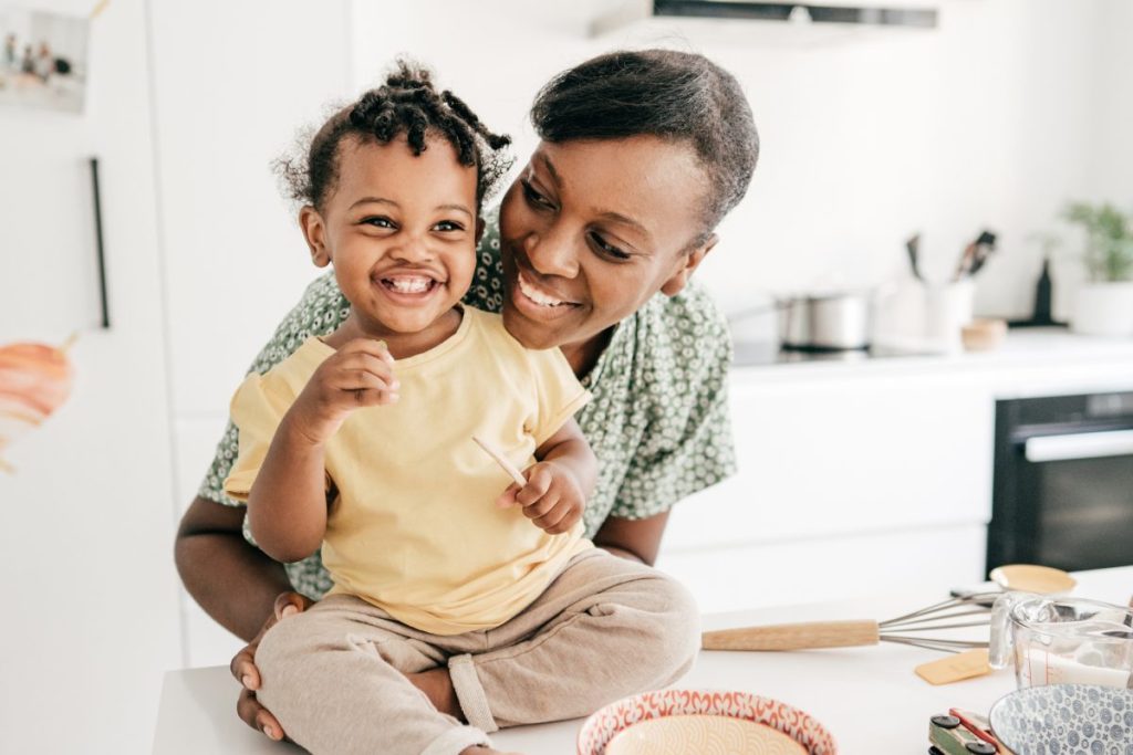 A toddler sitting on the counter while the parent hugs them with prepped food on the counter.