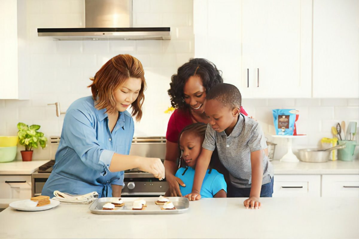 A woman showing another woman and her kids how to cook in the kitchen.