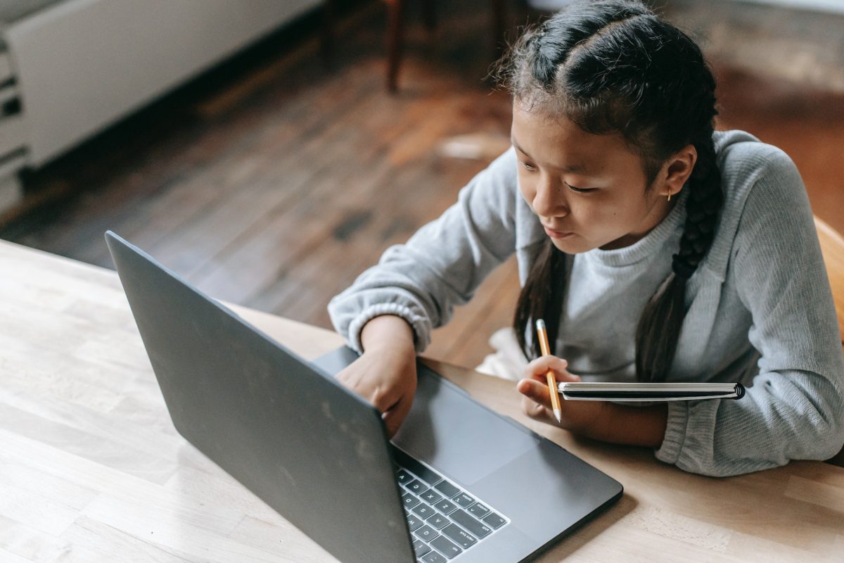A child sitting at a desk while holding a notebook and typing on the laptop.