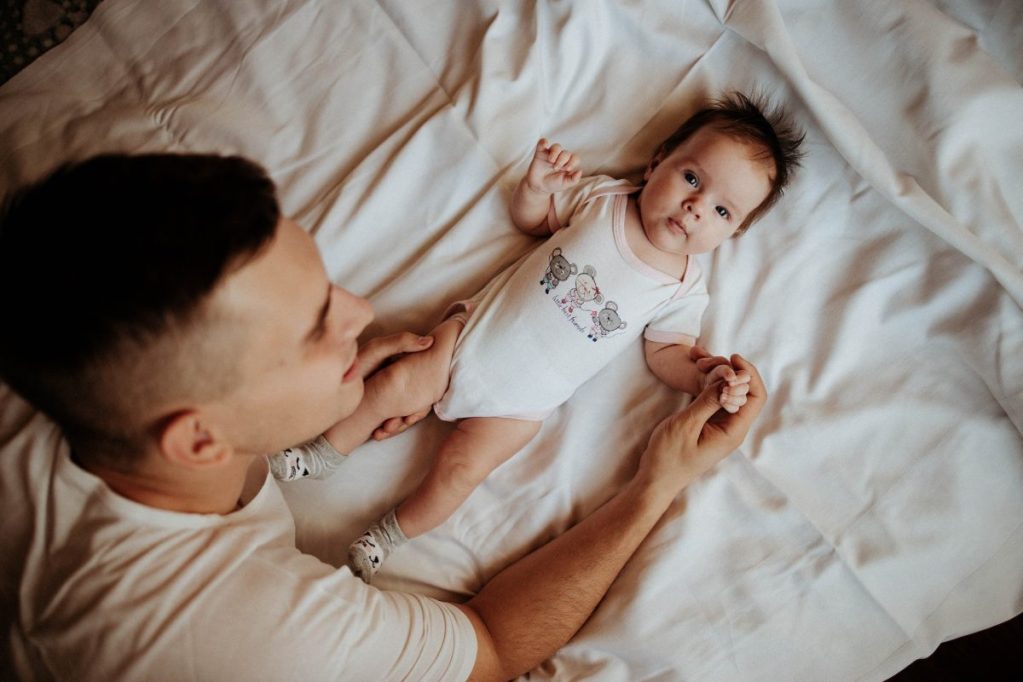 A parent looking at a baby laying down on a bed.