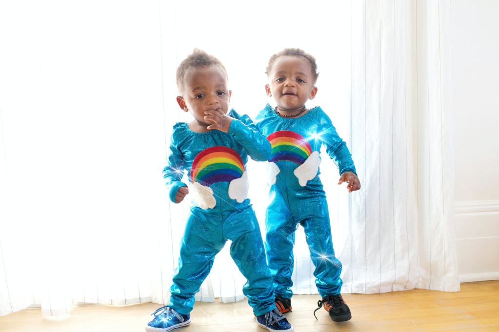 Twin babies wearing rainbow outfits standing up.