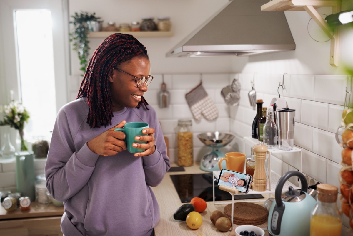 Smiling woman having coffee in kitchen using phone to monitor her sleeping baby